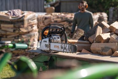 Person mit Gehörschutz steht neben einer Motorsäge und gestapeltem Holz.
