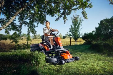 Mann mäht mit einem Husqvarna Rider Rasenmäher in einer grünen, sonnigen Landschaft.