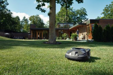 Ein Husqvarna Automower Mähroboter auf grünem Gras vor einem modernen Haus mit Bäumen.